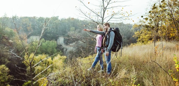 Tourists visiting surroundings from top of hill. Caucasian man with backpack points his hand into distance, charming blonde looks carefully. Traveling on foot concept.