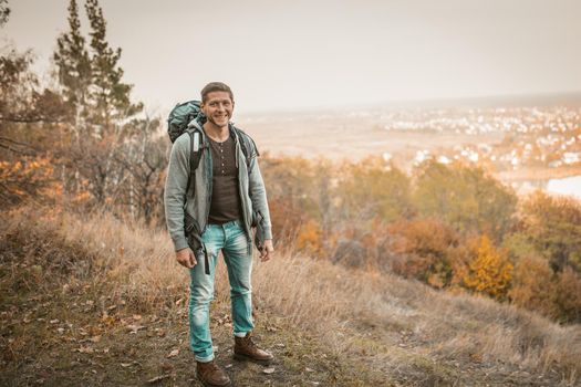 Traveler Stopped On Hillside, Young Caucasian Man With Large Backpack Smiles Looking At The Camera Against The Background Of Autumn Nature Below