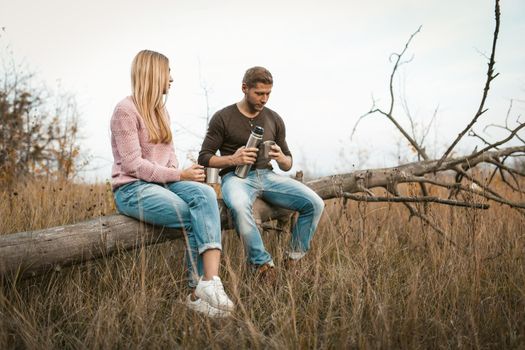 Coffee Break In Nature, Man And Woman Drink Hot Tea Pouring It From Thermos Into Camping Cups, Couple Sitting On Fallen Trunk Of Dried Tree Against Background Of Autumn Grass And Cloudy Sky