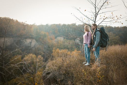 Couple of tourists admiring the sunset standing on the edge of a cliff holding hands, a Caucasian young man and woman standing in nature in the rays of the setting sun, side view. Hiking concept.