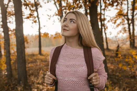 Traveling Woman Admires Beauty Of Autumn Forest Outdoors, Cheerful Blonde With Backpack Stands Looking At Side, With Autumn Yellow Trees On Sunny Blurred Back