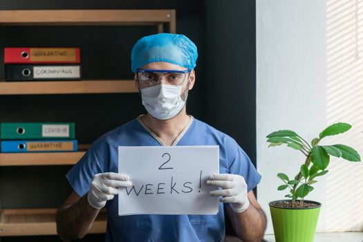 Male Doctor With Signs TWO WEEKS, Standing Near Window In Medical Uniform And Protective Mask, Doctor Shows Quarantine Countdown In Week Sheets, Healthcare Concept