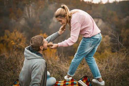 Loving Couple Has Break While Camping Outdoors, Man And Woman Feed Each Other Sandwiches While Sitting On Picnic Blanket Near Cliff Edge In Autumn Nature
