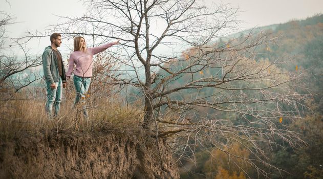 Couple Stands In Nature Holding Hands, A Man And A Woman Standing On The Edge Of A Cliff, Young Beautiful Girl With Long Hair Shows With Hand Into The Distance