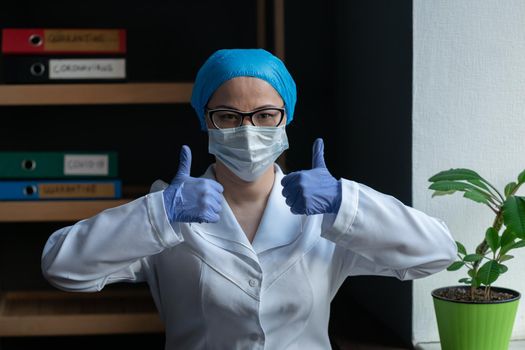 Female Doctor Showing Thumbs Up During Coronavirus Pandemic, Asian Woman Wearing Medical Uniform With Protective Mask And Eye Glasses Shows GREAT Hand Sign Standing Near Window In Office