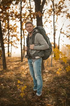 Hiking Of Male Backpacker In Autumn Nature Outdoors, Caucasian Man With Large Backpack Wearing Jeans And Gray Jacket Stopped In Background Of Autumn Forest Lit By Sun
