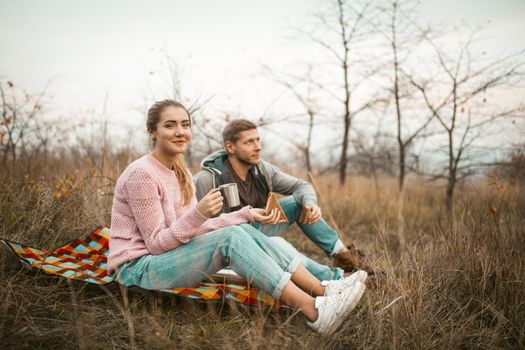 Young Men And Women Have Picnic Outdoors, Couple Of Travelers Eat Snacks And Drink A Hot Drink, Resting Sitting On Autumn Grass, Halt In Hiking Trip Concept