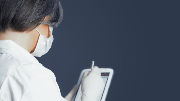 Health officer works with digital tablet. Gray-haired female doctor make notes in electronic database on digital gadget, rear or side view.