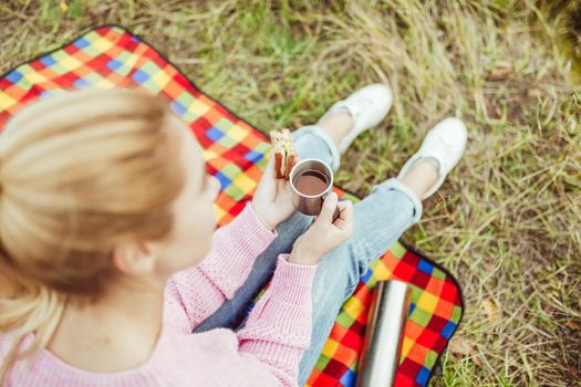 Charming blonde eats sandwich and drinks tea sitting on picnic blanket at grass. Focus on female hands. Close up shot, high angle view. Snack in nature outdoors concept.