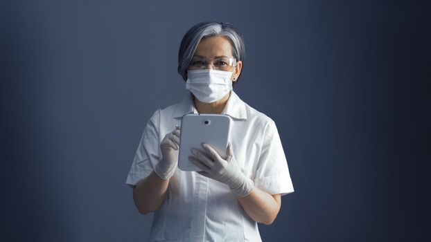 Female medic works with electronic notepad. Pretty woman in white uniform using apps in smart digital gadget in her work standing on blue gray background with Copy space on both sides. Toned image.