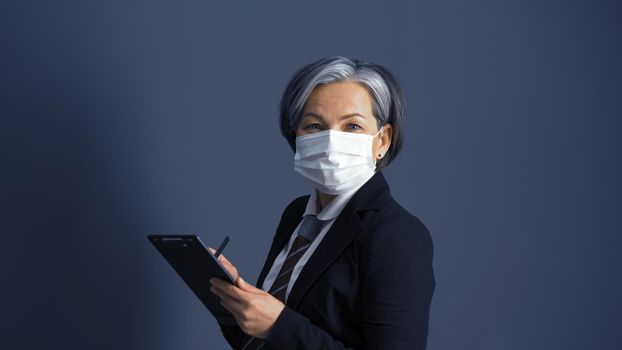 Gray haired businesswoman in protective mask smiles looking at camera on gray background. Pandemic concept.