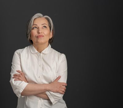 Creative mature woman looking upward thoughtfully with her arms crossed. Charming silver-haired woman smiling tenderly on dark grey background with textspace at right side.