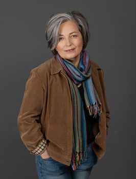 Pretty gray-haired woman in casual. Lovely mature lady wearing brown jacket and colorfull scarf standing hands in pockets on on gray background. Female studio portrait.