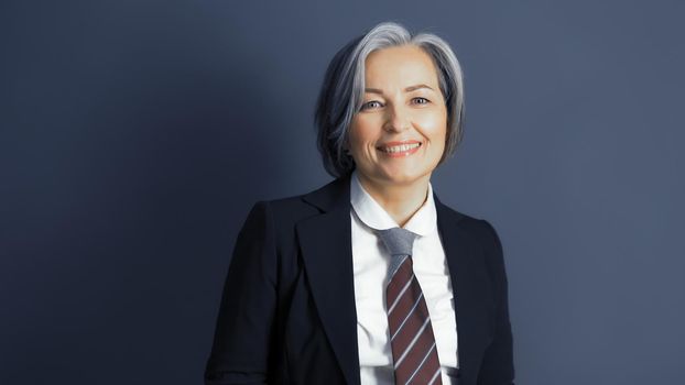 Positive middle-aged businesswoman without mask smiles happily. Optimistic graying woman wearing formalwear posing on blue gray background. Text or Copy space on both sides. Studio shot. Toned image.