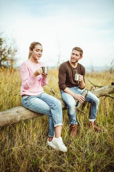 Tea party or coffee drinking outdoors. Cheerful couple drink hot coffee or tea communicate sitting on a wooden log outdoors.