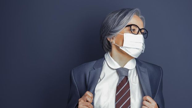 Mature woman wearing mask corrects her jacket looking at right side. Caucasian smart business woman on gray background. Copy space at left. Quarantine concept. Toned image.