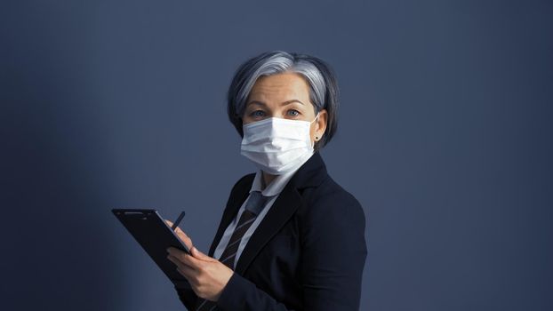 Confident middle-aged businesswoman in protective mask using digital notepad. Graying female model looks at camera, isolated on gray background in studio. Toned image.