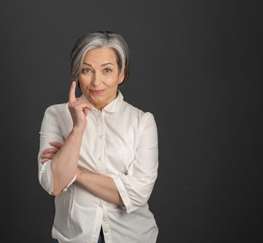 Have idea gesture from mature woman. Gray-haired businesswoman raised her index finger up while looking at camera posing on grey background Copyspace at right side. Eureka concept. Attention concept.