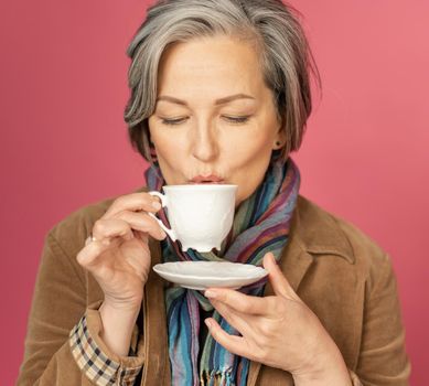 Coffee time concept. Charming Caucasian gray-haired woman drinks coffee in white cup on a pink background in studio. Close up shot.