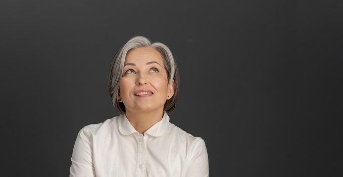 Thinking white-haired business woman looking upward. Creative middle-aged pretty woman thoughtfully smiles posing on grey background. Close up portrait. Copyspace at right side.