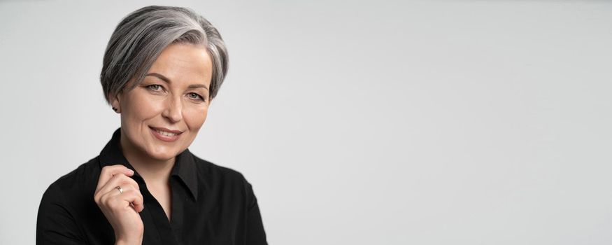 Pretty mature woman smiles with hand near face. Charming gray-haired lady in black blouse on white background. Copy spase for text. Horizontal blank or template for ad banner.