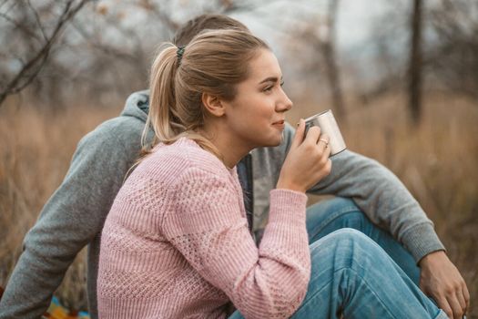 Picnic in nature. Young woman and man rests drinking hot coffee or tea while sitting on blanket at autumn grass outdoors. Profile view. Lifestyles concept.