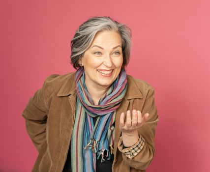 Funny mature woman laughs gesturing by hand and looking at side. Creative aged lady posing in studio on pink background.