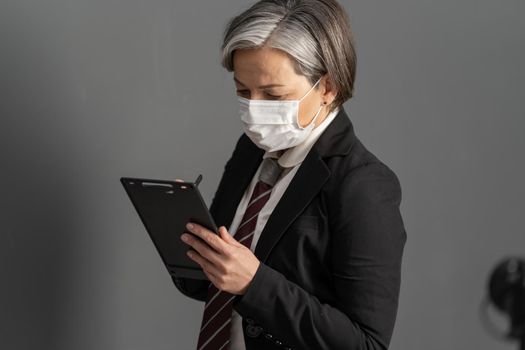 Caucasian businesswoman in protective mask works with notepad. Side view of a graying woman using digital gadget. Female office worker working during covid-19 pandemic. Close up portrait.