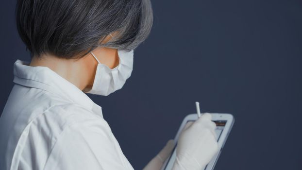 Pretty doctor in white uniform using apps in smart digital notepad in her work standing back on blue gray background. Text space on right. Rear view. Toned image.