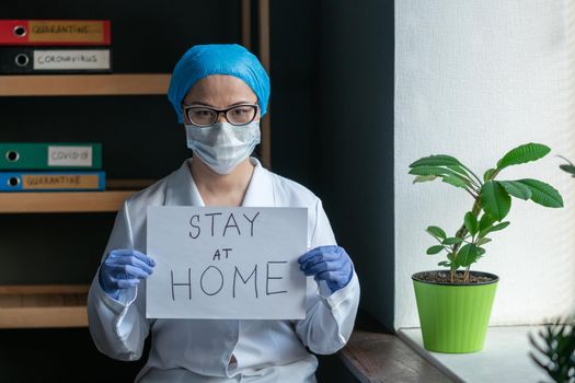 Doctor Shows Sign STAY AT HOME, Woman Wearing Protective Mask And White Medical Uniform Standing In The Office Next To The Window, Quarantine Concept