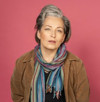 Sexy silver-haired woman looks at camera. Good-looking mature lady in multy-colored scarf posing in studio on pink background.