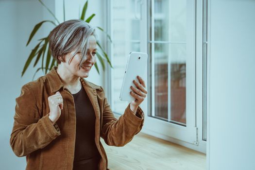 Beautiful mature woman communicates online on digital tablet. Silver-haired woman smiles talking while standing near window. Tinted image.