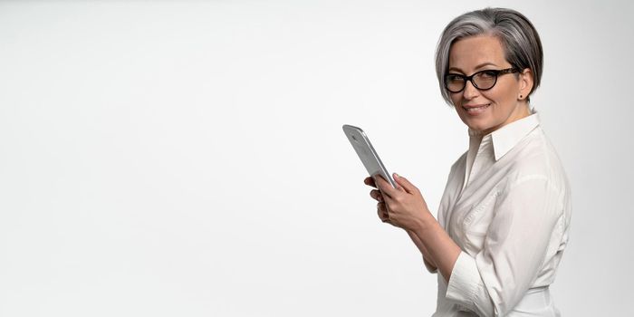 Cheerful business woman works on digital tablet on white background. Mature lady in white shirt looking at camera. Horizontal blank with empty spase for text at left side. Business or Education concept.