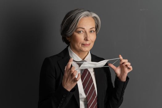 Charming gray-haired business woman putting on protective mask. Mature Caucasian woman wearing suit with tie looking at camera. Pandemic concept.