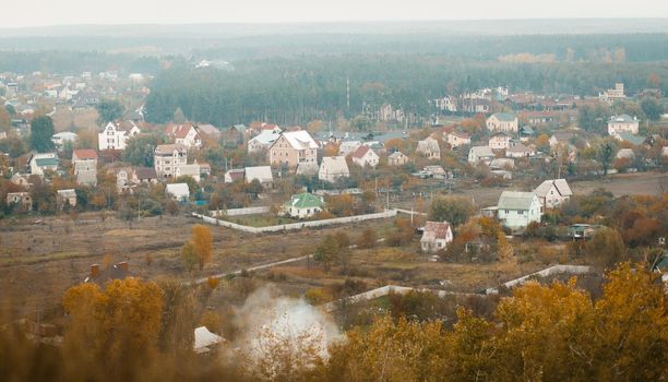 Panoramic view from the mountain to the cottage town, green lawns and coniferous forests on the horizon. Trees with colorful fall foliage in foreground. Autumnal picturesque landscape. Aerial View.