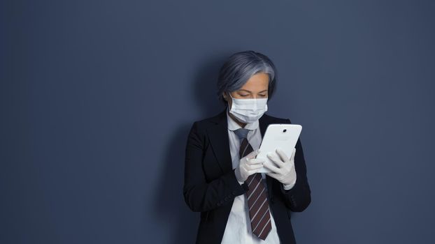 Mature woman works with digital tablet. Gray-haired businesswoman in black suit using electronic gadget standing against on gray office wall.