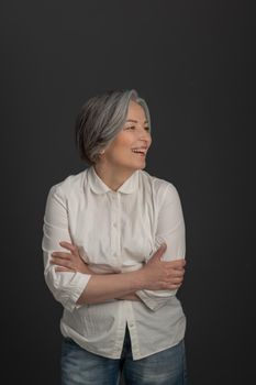 Laughing charming woman looks away confusedly with her arms folded. Joyful mature woman posing in studio on gray studio background.