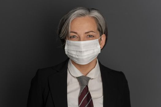 Pretty Caucasian businesswoman wearing protective mask and formalwear smiles on gray background. Copy space on both sides. Close up portrait. Outbreak coronavirus concept.