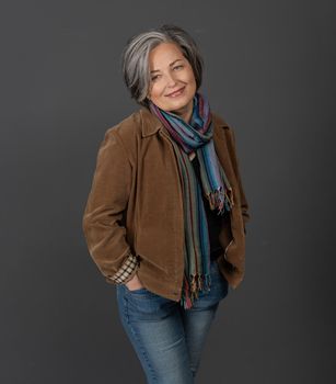 Pretty grey-haired woman in casual smiles posing on gray background. Elegant woman in jeans standing with his hands in his pockets.