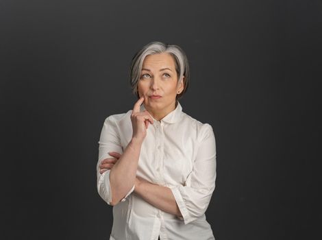 Serious gray-haired woman thinks looking up while touching her cheek with hand. Thoughtful middle-aged woman in white shirt cut out on gray back. Copy space on both sides.