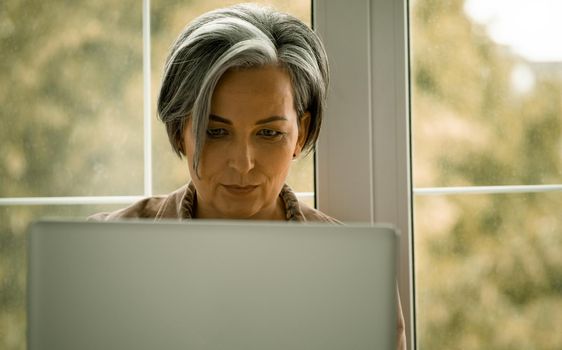 Mature woman works laptop sitting on a windowsill. Serious Caucasian Lady looking at computer monitor against window. Front view. Close up shot. Tinted image.