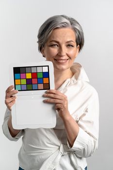 Mature woman holds color checker smiling at camera. Gray haired woman in white blouse shows target for color adjustment. Isolated on white background.