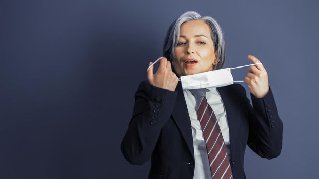 Business woman removes protective mask from her face. Focus on female face. Mature woman in formalwear on blue gray background. Copy space at left side. Close up portrait. Tinted image.