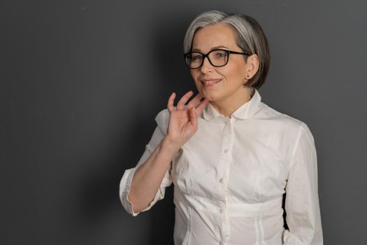 Graying pretty woman smiles touching chin with hand coquettishly. Smart mature lady in eyeglasses and white shirt on gray background. Text space at left side.