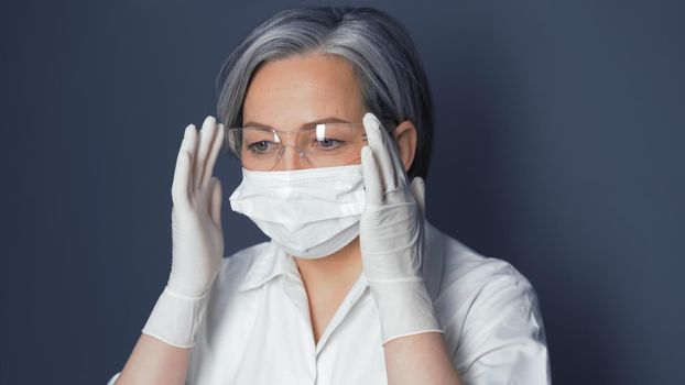 Doctor in uniform adjusts safety glasses. Cute Caucasian woman wearing protective mask and disposable gloves adjusts her glasses. Close up portrait. Toned image.