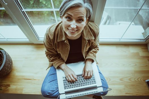 Smiling mature woman looks at camera typing laptop while sitting on a windowsill. High angle view. Tinted image.