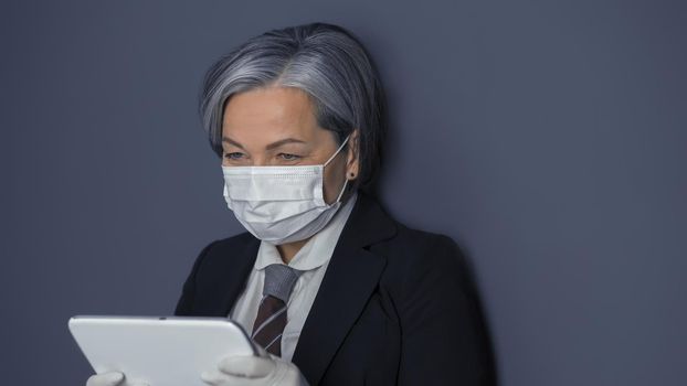 Mature businesswoman in protective mask using digital tablet. Gray haired woman reading news or working at tablet computer while standing on gray wall background. Toned image.