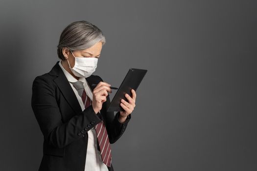 Caucasian businesswoman in protective mask working on tablet computer. Mature lady in formalwear using digital gadget. Empty space for text at right side. Quarantine concept.