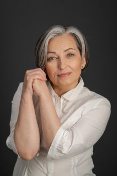 Aged woman charmingly smiles tilting head slightly and clasped hands near face. Beautiful lady in white shirt Isolated on gray.