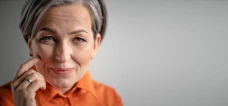 Charming gray-haired woman smiles touching cheek with finger. Selective focus on female face with tiny wrinkles. Close up portrait. Mature skin care concept. Horizontal blank witn copy spase for text.